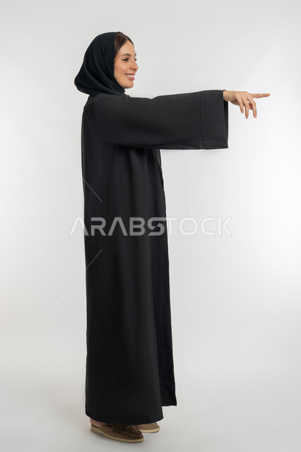 تحديد موقع شيء معين, التوضيح والدلالة لمكان محدد, بورتريه من الجانب لامرأة عربية خليجية إماراتية محجبة ترتدي العباءة السوداء تشير بإصبع السبابة للأمام، شابة سعودية تقوم بالإشارة إلى عنصر ما, الجسم بالطول الكامل، خلفية بيضاء