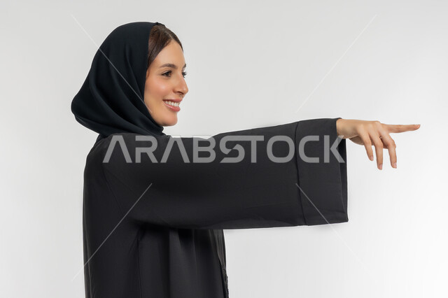 تحديد موقع شيء معين, التوضيح والدلالة لمكان محدد, بورتريه مقرب من الجانب لامرأة عربية خليجية إماراتية محجبة ترتدي العباءة السوداء تشير بإصبع السبابة للأمام، شابة سعودية تقوم بالإشارة إلى عنصر ما, خلفية بيضاء