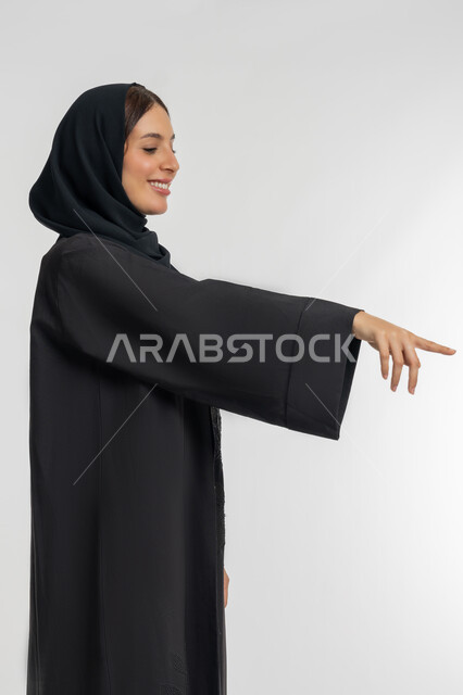 التوضيح والدلالة لمكان محدد, الإشارة إلى عنصر ما, بورتريه من الجانب لامرأة عربية خليجية إماراتية محجبة ترتدي العباءة السوداء تشير بإصبع السبابة للأسفل، شابة سعودية تقوم بتحديد موقع شيء معين, خلفية بيضاء