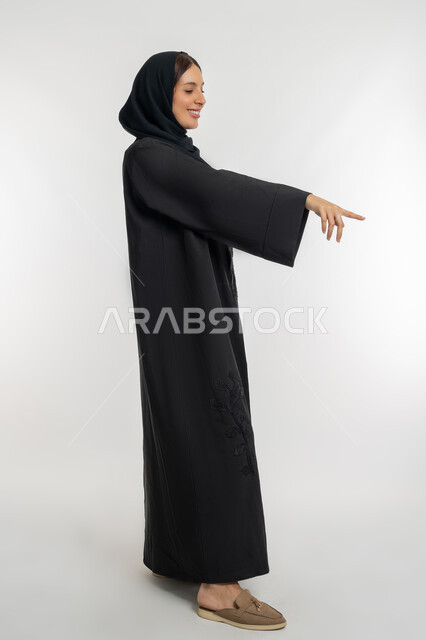 التوضيح والدلالة لمكان محدد, الإشارة إلى عنصر ما, بورتريه من الجانب لامرأة عربية خليجية إماراتية محجبة ترتدي العباءة السوداء تشير بإصبع السبابة للأسفل، شابة سعودية تقوم بتحديد موقع شيء معين, الجسم بالطول الكامل, خلفية بيضاء