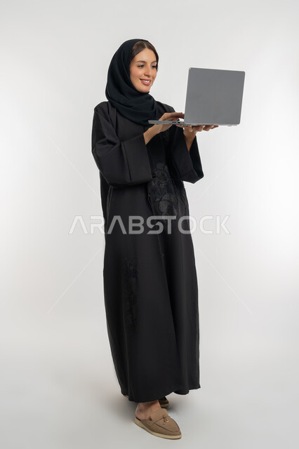 مفهوم العمل عن بعد, استخدام الأجهزة التقنية الحديثة والمتطورة,  بورتريه لامرأة عربية خليجية إماراتية محجبة ترتدي العباءة السوداء تعمل على جهاز الحاسوب المحمول, شابة سعودية تقوم بإنجاز المهام, الجسم بالطول الكامل، خلفية بيضاء