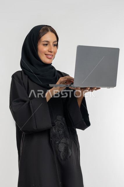 استخدام الأجهزة التقنية الحديثة والمتطورة, مفهوم العمل عن بعد, بورتريه لامرأة عربية خليجية إماراتية محجبة ترتدي العباءة السوداء تعمل على جهاز الحاسوب المحمول, شابة سعودية تقوم بإنجاز المهام, خلفية بيضاء