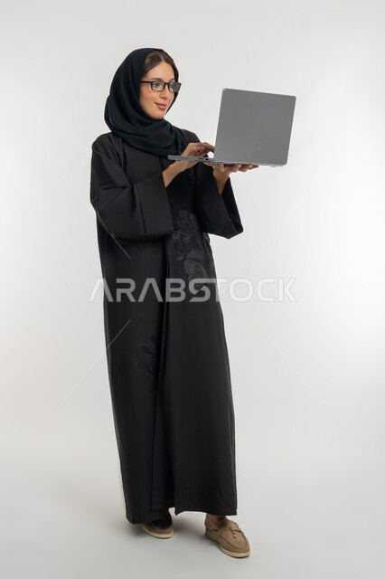 Remote work concept, use of modern and advanced technical devices, portrait of a veiled Emirati Gulf Arab woman wearing a black abaya working on a laptop, a young Saudi woman completing tasks, full-length body, white background
