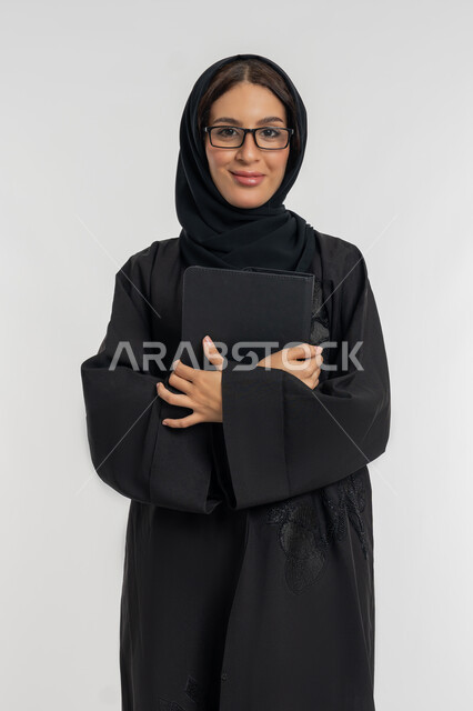شابة سعودية مبتسمة تحمل التابلت, دمج التقنيات الحديثة والتكنولوجيا في الحياة العملية, بورتريه لامرأة عربية خليجية إماراتية محجبة ترتدي العباءة تضع نظارة طبية تمسك بيدها الجهاز اللوحي, خلفية بيضاء