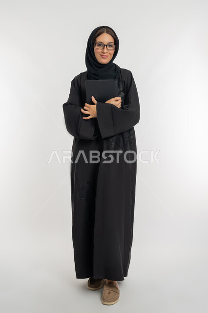 دمج التقنيات الحديثة والتكنولوجيا في الحياة العملية, بورتريه لامرأة عربية خليجية إماراتية محجبة ترتدي العباءة تضع نظارة طبية تمسك بيدها الجهاز اللوحي, شابة سعودية مبتسمة تحمل التابلت, صورة الجسم بالطول الكامل، خلفية بيضاء
