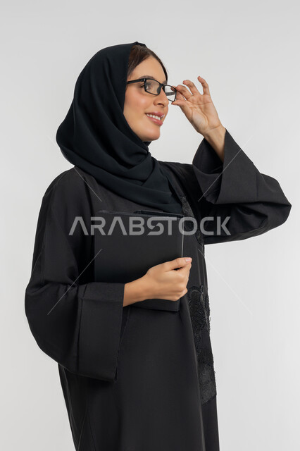 شابة سعودية مبتسمة تحمل التابلت, دمج التقنيات الحديثة والتكنولوجيا في الحياة العملية, بورتريه لامرأة عربية خليجية إماراتية محجبة ترتدي العباءة تضع نظارة طبية تمسك بيدها الجهاز اللوحي, خلفية بيضاء