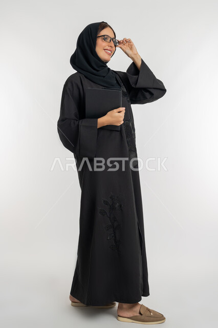 Integrating modern technologies and technology into practical life, portrait of a veiled Gulf Arab Emirati woman wearing an abaya, wearing glasses and holding a tablet, smiling young Saudi woman holding a tablet, full body image, white background