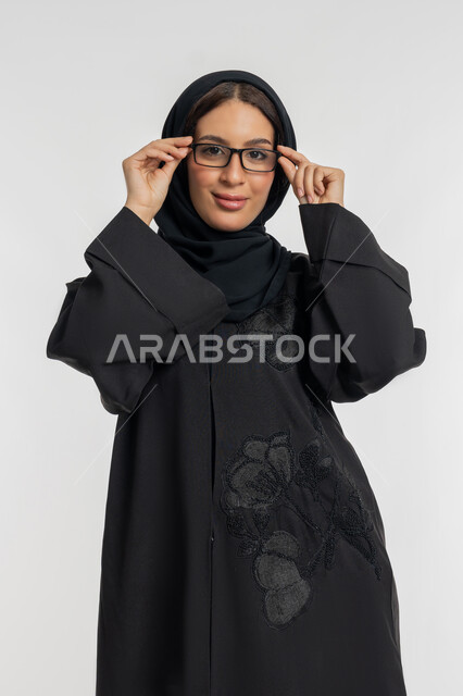 مفهوم العناية بالصحة والمظهر الخارجي، ضعف وقصر النظر، الاهتمام بالموضة والأناقة, بورتريه لامرأة عربية خليجية إماراتية ترتدي العباءة والحجاب تمسك بيدها النظارات الطبية بتعابير الثقة بالنفس, شابة سعودية تنظر الى الكاميرا بإيماءات السرور, خلفية بيضاء