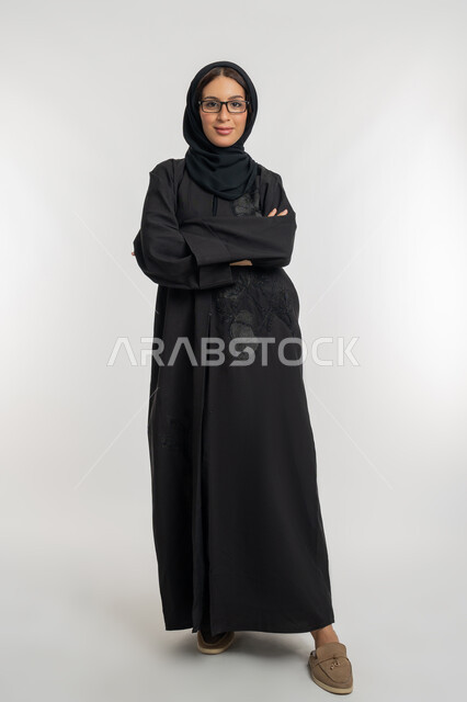 Myopia and shortsightedness, interest in fashion and elegance, concept of health care, portrait of an Arab Gulf Emirati woman wearing an abaya and hijab, wearing glasses, standing with crossed hands with an expression of self-confidence, a young Saudi woman looking at the camera with gestures of pleasure, full-length body, white background
