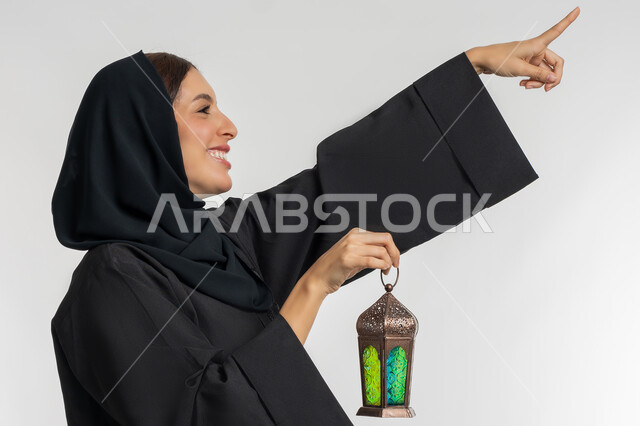 البهجة بقدوم شهر الخير, الاحتفال بالمناسبات الدينية الإسلامية, التحضير لاستقبال رمضان المبارك, بورتريه من الجانب لامرأة عربية خليجية إماراتية محجبة ترتدي العباءة السوداء تمسك بيدها فانوس معدني مزخرف, شابة سعودية تشير بإصبع السبابة إلى شيء ما, خلفية بيضاء