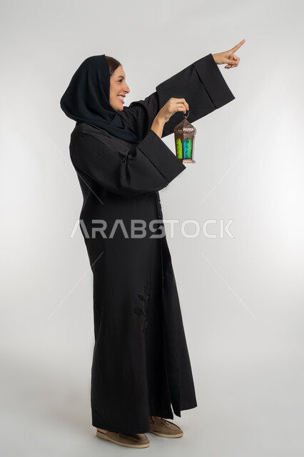Joy of the arrival of the month of goodness, celebration of Islamic religious occasions, preparation for the reception of the holy month of Ramadan, side portrait of a veiled Emirati Gulf Arab woman wearing a black abaya holding a decorative metal lantern in her hand, a young Saudi woman pointing with her index finger at something, white background