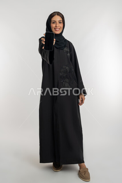 Mobile application display, e-marketing using technology and technology, portrait of a smiling Emirati Gulf Arab woman wearing hijab and abaya holding a mobile phone with a blank black screen, a young Saudi woman looking at the camera with an expression of pleasure, full body, white background