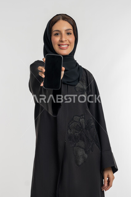 عرض تطبيقات الهاتف المحمول، التسويق الالكتروني باستخدام التقنية والتكنولوجيا، بورتريه لإمراة عربية خليجية اماراتية مبتسمة ترتدي الحجاب والعباءة تحمل بيدها جوال بشاشة سوداء فارغة، شابة سعودية تنظر الى الكاميرا بتعابير السرور، خلفية بيضاء