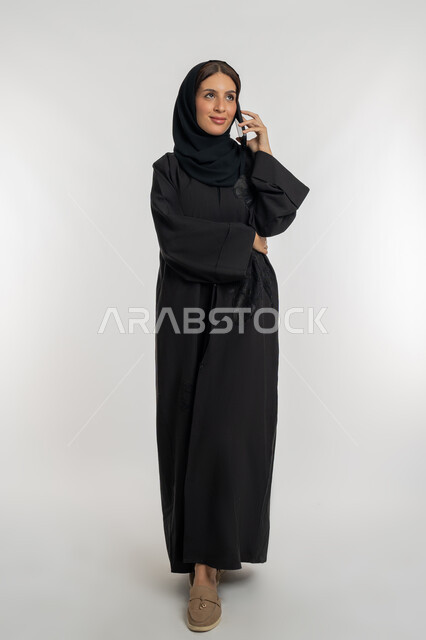 التواصل مع العائلة والأصدقاء, استخدام الأجهزة التقنية الحديثة في الحياة اليومية, مشاركة الأخبار والأحاديث الجيدة, بورتريه لامرأة عربية خليجية إماراتية محجبة ترتدي العباءة السوداء تمسك بيدها الهاتف المحمول, شابة سعودية تقوم بإجراء اتصال هاتفي, خلفية بيضاء