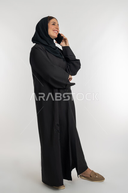 Communicating with family and friends, using modern technology devices in daily life, sharing good news and conversations, portrait of a veiled Emirati Gulf Arab woman wearing a black abaya holding a mobile phone in her hand, a young Saudi woman making a phone call, white background