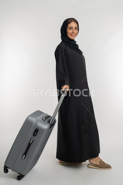 التجهيز للسفرات الممتعة, الاستمتاع بالرحلات السياحية في وقت العطلة, بورتريه لامرأة عربية خليجية إماراتية محجبة ترتدي العباءة السوداء تمسك بيدها حقيبة سفر رمادية, شابة سعودية تنظر إلى الكاميرا بإيماءات السعادة والسرور، الجسم بالطول الكامل, خلفية بيضاء