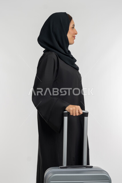 الاستمتاع بالرحلات السياحية في وقت العطلة, التجهيز للسفرات الممتعة, بورتريه من الجانب لامرأة عربية خليجية إماراتية محجبة ترتدي العباءة السوداء تمسك بيدها حقيبة سفر رمادية, شابة سعودية تنظر إلى شيء ما بإيماءات السعادة والسرور، خلفية بيضاء