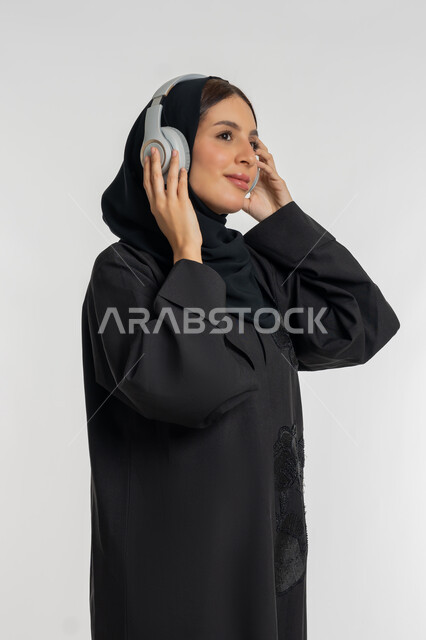 تعابير الاندماج و التفاعل مع الأنغام الموسيقية, استخدام تقنية الهيدفون الحديثة, الاستمتاع بسماع الأغاني المفضلة, بورتريه لامرأة عربية خليجية إماراتية محجبة ترتدي العباءة السوداء تضع سماعات الرأس الصوتية, شابة سعودية تستمع إلى الموسيقا, خلفية بيضاء