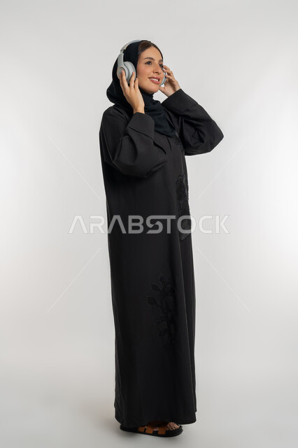 استخدام تقنية الهيدفون الحديثة, تعابير الاندماج و التفاعل مع الأنغام الموسيقية, الاستمتاع بسماع الأغاني المفضلة, بورتريه لامرأة عربية خليجية إماراتية محجبة ترتدي العباءة السوداء تضع سماعات الرأس الصوتية, شابة سعودية تستمع إلى الموسيقا، الجسم بالطول الكامل، خلفية بيضاء