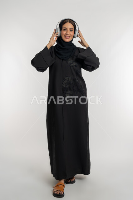 استخدام تقنية الهيدفون الحديثة, تعابير الاندماج و التفاعل مع الأنغام الموسيقية, الاستمتاع بسماع الأغاني المفضلة, بورتريه لامرأة عربية خليجية إماراتية محجبة ترتدي العباءة السوداء تضع سماعات الرأس الصوتية, شابة سعودية تستمع إلى الموسيقا، الجسم بالطول الكامل، خلفية بيضاء