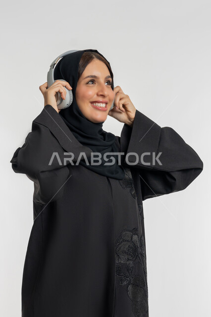 تعابير الاندماج و التفاعل مع الأنغام الموسيقية, استخدام تقنية الهيدفون الحديثة, الاستمتاع بسماع الأغاني المفضلة, بورتريه لامرأة عربية خليجية إماراتية محجبة ترتدي العباءة السوداء تضع سماعات الرأس الصوتية, شابة سعودية تستمع إلى الموسيقا, خلفية بيضاء