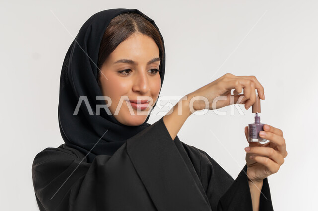 مفهوم العناية بالأظافر, الاهتمام بالأناقة والمظهر الخارجي, التسويق لمنتجات ومستحضرات التجميل, بورتريه مقرب من الجانب لامرأة عربية خليجية إماراتية محجبة ترتدي العباءة تمسك بيدها علبة طلاء الأظافر, شابة سعودية تقوم بالتزين والتجمل, خلفية بيضاء