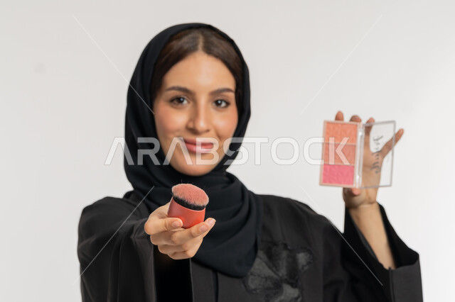استخدام المساحيق التجميلية, العناية بالمظهر الخارجي, التسويق لمستحضرات ومنتجات التجميل, مفهوم التزين والتجمل, بورتريه مقرب لامرأة عربية خليجية إماراتية محجبة ترتدي العباءة السوداء تستعمل فرشاة أحمر الخدود, شابة سعودية تضع المكياج, خلفية بيضاء