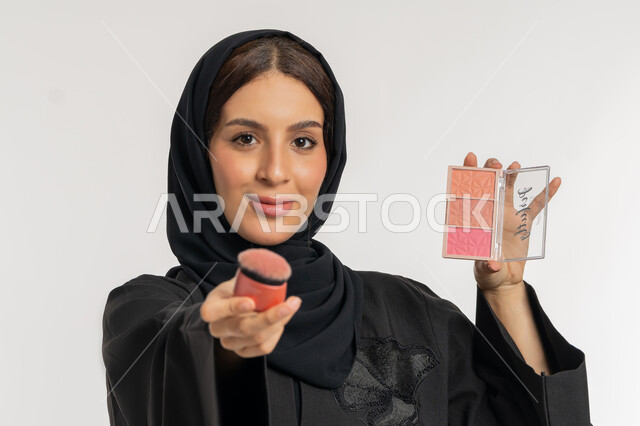 استخدام المساحيق التجميلية, العناية بالمظهر الخارجي, التسويق لمستحضرات ومنتجات التجميل, مفهوم التزين والتجمل, بورتريه مقرب لامرأة عربية خليجية إماراتية محجبة ترتدي العباءة السوداء تستعمل فرشاة أحمر الخدود, شابة سعودية تضع المكياج, خلفية بيضاء