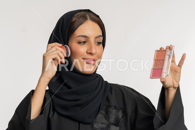 العناية بالمظهر الخارجي, التسويق لمستحضرات ومنتجات التجميل, استخدام المساحيق التجميلية, مفهوم التزين والتجمل, بورتريه مقرب لامرأة عربية خليجية إماراتية محجبة ترتدي العباءة السوداء تستعمل فرشاة أحمر الخدود, شابة سعودية تضع المكياج, خلفية بيضاء