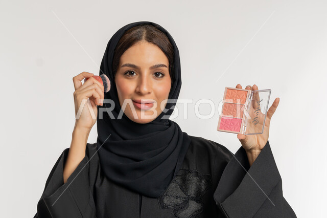 الاهتمام والعناية بالمظهر الخارجي, التسويق لمستحضرات ومنتجات التجميل, استخدام المساحيق التجميلية, مفهوم التزين والتجمل, بورتريه مقرب لامرأة عربية خليجية إماراتية محجبة ترتدي العباءة السوداء تستعمل فرشاة أحمر الخدود, شابة سعودية تضع المكياج, خلفية بيضاء