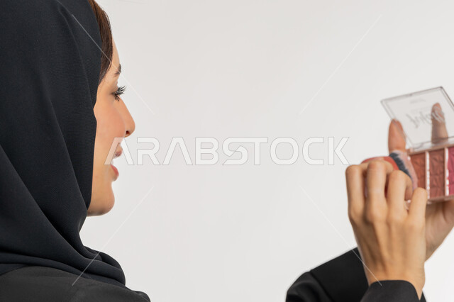 التسويق لمستحضرات ومنتجات التجميل, استخدام المساحيق التجميلية, الاهتمام والعناية بالمظهر الخارجي, مفهوم التزين والتجمل, بورتريه مقرب من الخلف لامرأة عربية خليجية إماراتية محجبة ترتدي العباءة السوداء تستعمل فرشاة أحمر الخدود, شابة سعودية تضع المكياج, خلفية بيضاء