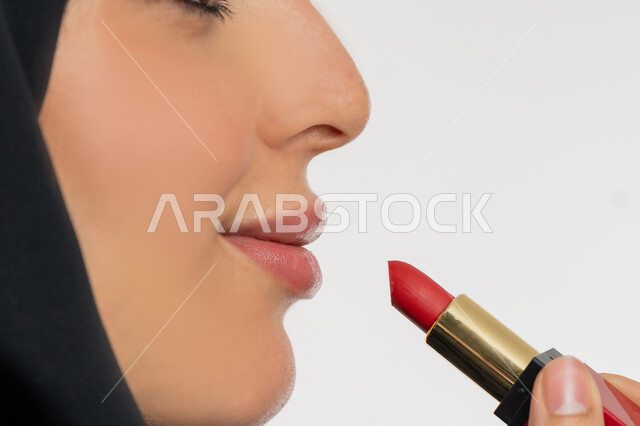 الترويج لمنتجات و مساحيق التجميل, استعمال المستحضرات التجميلية, مفهوم التزين والتجمل, العناية بالجمال والمظهر الخارجي, بورتريه مقرب من الجانب لامرأة عربية خليجية إماراتية محجبة ترتدي العباءة السوداء تضع أحمر شفاه, شابة سعودية تقوم بوضع المكياج, خلفية بيضاء
