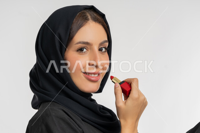 استعمال المستحضرات التجميلية,  مفهوم التزين والتجمل, الترويج لمنتجات و مساحيق التجميل, العناية بالجمال والمظهر الخارجي, بورتريه مقرب من الجانب لامرأة عربية خليجية إماراتية محجبة ترتدي العباءة السوداء تضع أحمر شفاه, شابة سعودية تقوم بوضع المكياج, خلفية بيضاء