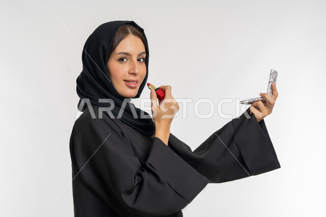 الترويج لمنتجات و مساحيق التجميل, استعمال المستحضرات التجميلية, مفهوم التزين والتجمل, العناية بالجمال والمظهر الخارجي, بورتريه مقرب من الجانب لامرأة عربية خليجية إماراتية محجبة ترتدي العباءة السوداء تضع أحمر شفاه, شابة سعودية تقوم بوضع المكياج, خلفية بيضاء