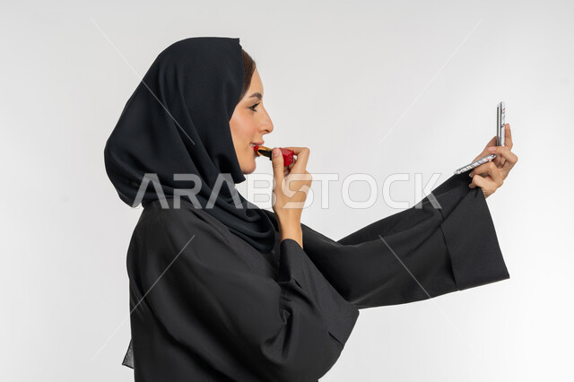 مفهوم التزين والتجمل, الترويج لمنتجات و مساحيق التجميل, استعمال المستحضرات التجميلية, العناية بالجمال والمظهر الخارجي, بورتريه مقرب من الجانب لامرأة عربية خليجية إماراتية محجبة ترتدي العباءة السوداء تضع أحمر شفاه, شابة سعودية تقوم بوضع المكياج, خلفية بيضاء