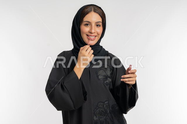 مفهوم التزين والتجمل, استعمال المستحضرات التجميلية, الترويج لمنتجات و مساحيق التجميل, العناية بالجمال والمظهر الخارجي, بورتريه مقرب لامرأة عربية خليجية إماراتية محجبة ترتدي العباءة الحمراء تضع أحمر شفاه, شابة سعودية تقوم بوضع الليب غلوس, خلفية بيضاء