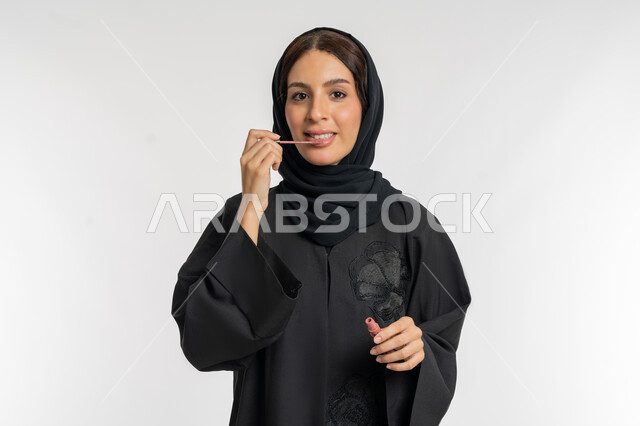 استعمال المستحضرات التجميلية, مفهوم التزين والتجمل, الترويج لمنتجات و مساحيق التجميل, العناية بالجمال والمظهر الخارجي, بورتريه مقرب لامرأة عربية خليجية إماراتية محجبة ترتدي العباءة الحمراء تضع أحمر شفاه, شابة سعودية تقوم بوضع الليب غلوس, خلفية بيضاء
