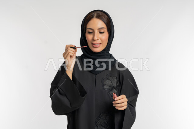 مفهوم التزين والتجمل, استعمال المستحضرات التجميلية, الترويج لمنتجات و مساحيق التجميل, العناية بالجمال والمظهر الخارجي, بورتريه مقرب لامرأة عربية خليجية إماراتية محجبة ترتدي العباءة الحمراء تضع أحمر شفاه, شابة سعودية تقوم بوضع الليب غلوس, خلفية بيضاء