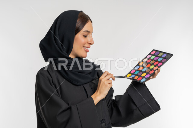 الاهتمام بالمظهر الخارجي, استخدام المستحضرات التجميلية, الترويج لمنتجات و مساحيق التجميل, مفهوم العناية بالجمال والبشرة, بورتريه مقرب لامرأة عربية خليجية إماراتية محجبة ترتدي العباءة السوداء تستعمل فرشاة ظل العيون, شابة سعودية تقوم بوضع المكياج, خلفية بيضاء