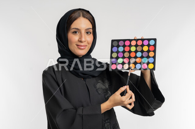 استخدام المستحضرات التجميلية, الترويج لمنتجات و مساحيق التجميل, الاهتمام بالمظهر الخارجي, مفهوم العناية بالجمال والبشرة, بورتريه مقرب لامرأة عربية خليجية إماراتية محجبة ترتدي العباءة السوداء تستعمل فرشاة ظل العيون, شابة سعودية تقوم بوضع المكياج, خلفية بيضاء