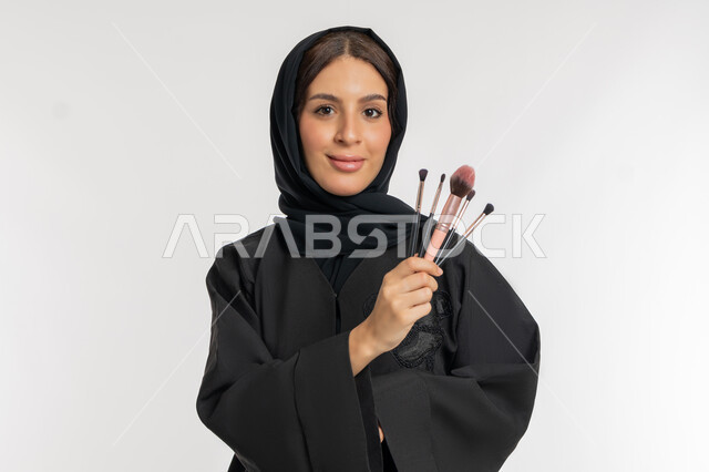 استخدام ادوات المستحضرات التجميلية، الترويج لمنتجات و مساحيق التجميل, مفهوم العناية بالجمال والبشرة, بورتريه مقرب لامرأة عربية خليجية إماراتية محجبة ترتدي العباءة السوداء تمسك بيدها الفراشي, شابة سعودية تنظر إلى الكاميرا بإيماءات السعادة والسرور, خلفية بيضاء