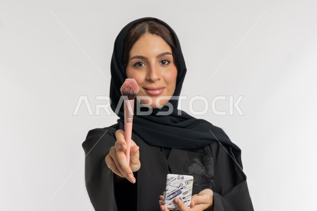 الاهتمام والعناية بالمظهر الخارجي, التسويق لمستحضرات ومنتجات التجميل, استخدام المساحيق التجميلية, مفهوم التزين والتجمل, بورتريه مقرب لامرأة عربية خليجية إماراتية محجبة ترتدي العباءة السوداء تستعمل فرشاة أحمر الخدود, شابة سعودية تضع المكياج, خلفية بيضاء