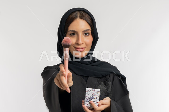 التسويق لمستحضرات ومنتجات التجميل, استخدام المساحيق التجميلية, الاهتمام والعناية بالمظهر الخارجي, مفهوم التزين والتجمل, بورتريه مقرب لامرأة عربية خليجية إماراتية محجبة ترتدي العباءة السوداء تستعمل فرشاة أحمر الخدود, شابة سعودية تضع المكياج, خلفية بيضاء