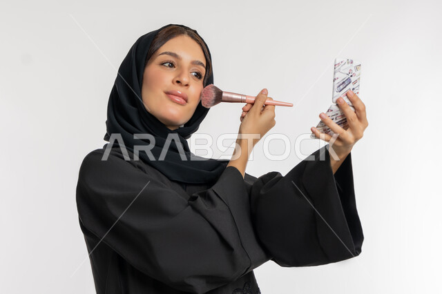 العناية بالمظهر الخارجي, التسويق لمستحضرات ومنتجات التجميل, استخدام المساحيق التجميلية, مفهوم التزين والتجمل, بورتريه مقرب لامرأة عربية خليجية إماراتية محجبة ترتدي العباءة السوداء تستعمل فرشاة أحمر الخدود, شابة سعودية تضع المكياج, خلفية بيضاء