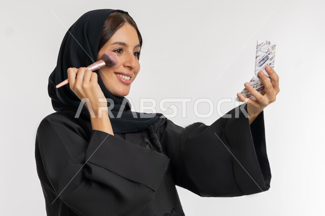 العناية بالمظهر الخارجي, التسويق لمستحضرات ومنتجات التجميل, استخدام المساحيق التجميلية, مفهوم التزين والتجمل, بورتريه مقرب لامرأة عربية خليجية إماراتية محجبة ترتدي العباءة السوداء تستعمل فرشاة أحمر الخدود, شابة سعودية تضع المكياج, خلفية بيضاء