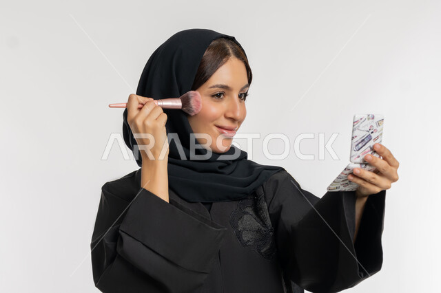 الاهتمام والعناية بالمظهر الخارجي, التسويق لمستحضرات ومنتجات التجميل, استخدام المساحيق التجميلية, مفهوم التزين والتجمل, بورتريه مقرب لامرأة عربية خليجية إماراتية محجبة ترتدي العباءة السوداء تستعمل فرشاة أحمر الخدود, شابة سعودية تضع المكياج, خلفية بيضاء