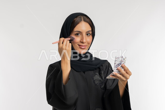 التسويق لمستحضرات ومنتجات التجميل, استخدام المساحيق التجميلية, الاهتمام والعناية بالمظهر الخارجي, مفهوم التزين والتجمل, بورتريه مقرب لامرأة عربية خليجية إماراتية محجبة ترتدي العباءة السوداء تستعمل فرشاة أحمر الخدود, شابة سعودية تضع المكياج, خلفية بيضاء