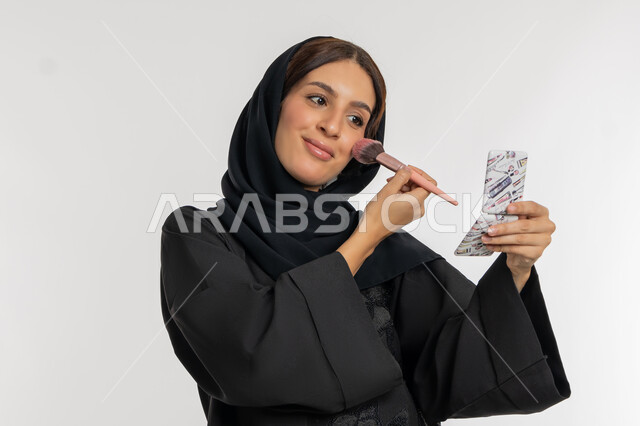 الاهتمام والعناية بالمظهر الخارجي, التسويق لمستحضرات ومنتجات التجميل, استخدام المساحيق التجميلية, مفهوم التزين والتجمل, بورتريه مقرب لامرأة عربية خليجية إماراتية محجبة ترتدي العباءة السوداء تستعمل فرشاة أحمر الخدود, شابة سعودية تضع المكياج, خلفية بيضاء