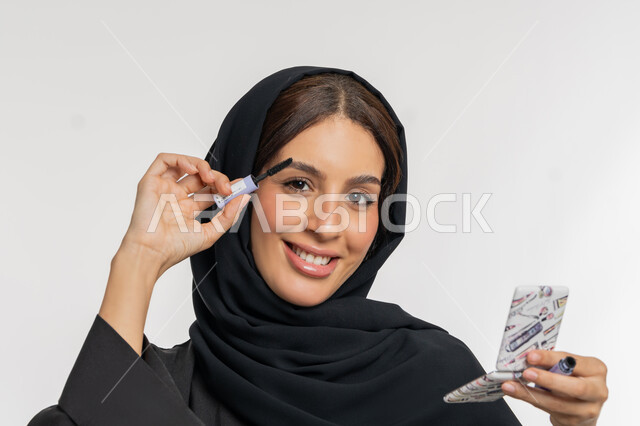 الترويج لمنتجات ومستحضرات التجميل, الاهتمام بالجمال والمظهر الخارجي, مفهوم التزين والتجمل, بورتريه مقرب لامرأة عربية خليجية إماراتية محجبة ترتدي العباءة السوداء تقوم باستخدام المسكرة, شابة سعودية تضع المكياج, خلفية بيضاء