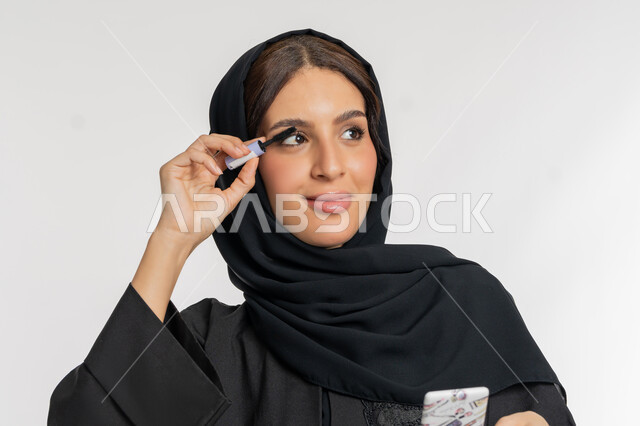 مفهوم التزين والتجمل, الترويج لمنتجات ومستحضرات التجميل, الاهتمام بالجمال والمظهر الخارجي,  بورتريه مقرب لامرأة عربية خليجية إماراتية محجبة ترتدي العباءة السوداء تقوم باستخدام المسكرة, شابة سعودية تضع المكياج, خلفية بيضاء